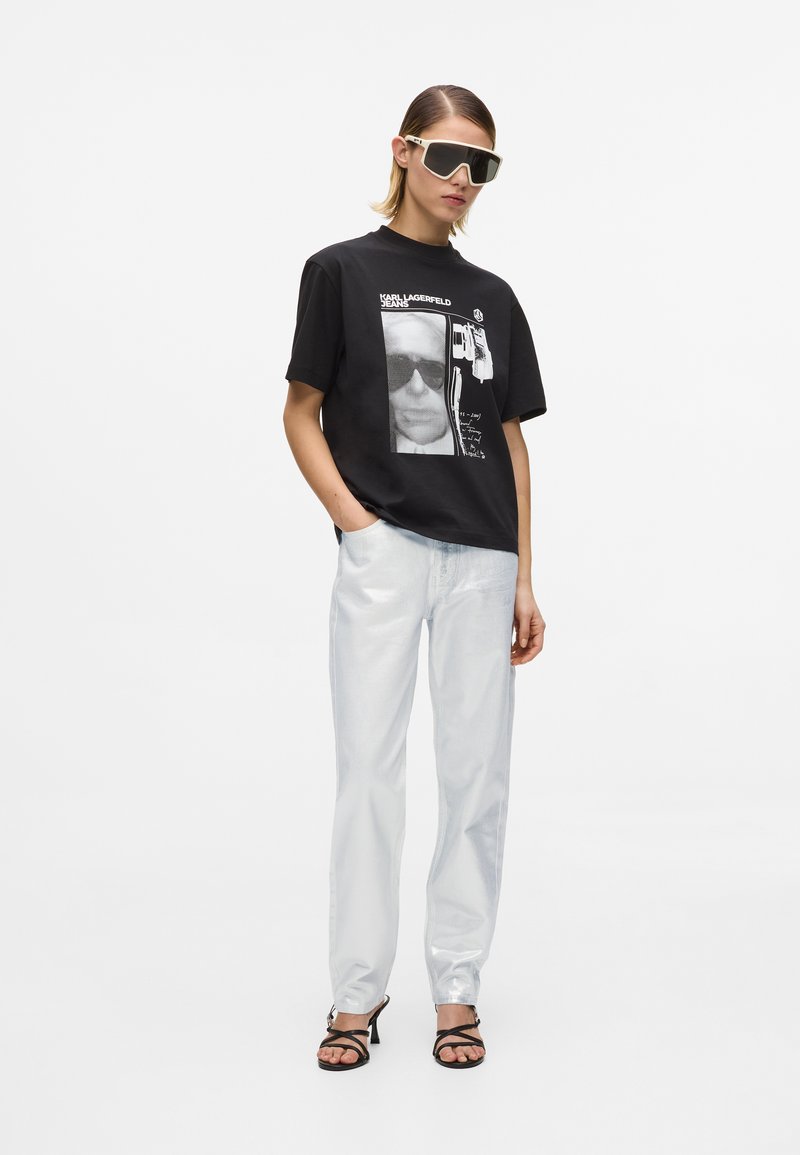 Γυναίκα που φοράει μαύρο γραφικό μπλουζάκι, ανοιχτό μπλε τζιν, μαύρα πέδιλα με τακούνι και μεγάλα λευκά γυαλιά ηλίου, στέκεται με το ένα χέρι στην τσέπη.