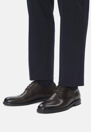 Ciemnobrązowe, wypolerowane skórzane buty wizytowe z czarnymi podeszwami, noszone z granatowymi spodniami i czarnymi skarpetkami na białym tle.