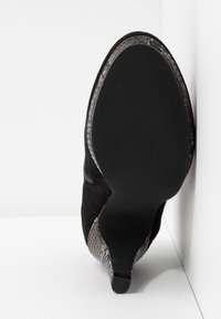 Bottine noire à talon avec semelle texturée à motif serpent, bout pointu et talon fin, positionnée contre des murs d'angle blancs.