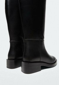 Bottes noires en cuir montant jusqu'au genou, avec un bout arrondi, des coutures minimalistes et un talon épais. Texture lisse avec un design épuré et moderne.