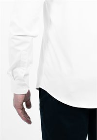 Witte lange mouwen shirt met een knoopsluiting bij de manchetten, met een zichtbaar logo op de mouw en een ronde zoom, gecombineerd met donkere broeken.