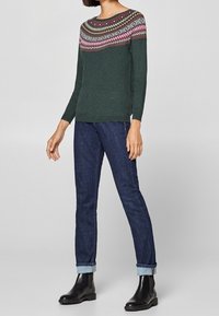 Maglione verde scuro con un collo decorato in tonalità di rosa, beige e bianco. Abbinato a jeans blu scuro attillati e stivaletti neri alla caviglia.