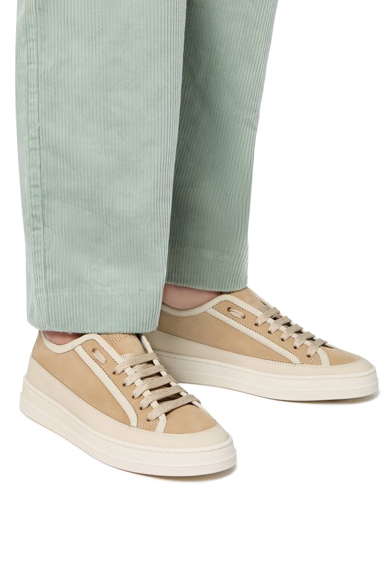 Des baskets beiges en cuir lisse avec des accents en daim, une semelle blanche contrastante et un design classique à lacets. Portées avec un pantalon en velours côtelé vert clair.