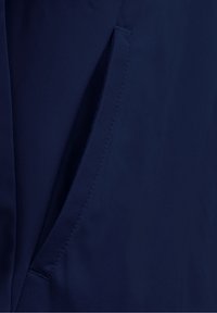 Primo piano di una tasca di una giacca blu navy con cuciture visibili e un'apertura obliqua su tessuto liscio.