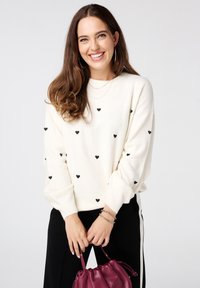 Crèmekleurige trui met zwarte hartpatronen, ronde halslijn en lange mouwen. Gecombineerd met zwarte broeken en een bordeauxrode handtas. Zilveren armbanden gedragen.