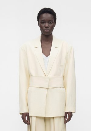 Femme aux cheveux courts et bouclés portant un blazer crème oversize avec une large ceinture et un pantalon plissé assorti, debout devant un fond blanc uni.