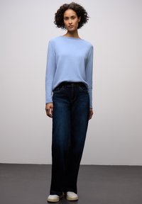 Jasnoniebieski sweter z szerokim dekoltem typu łódka, zestawiony z ciemnoniebieskimi szerokimi jeansami. Białe trampki dopełniają casualowy strój na jednolitym tle.