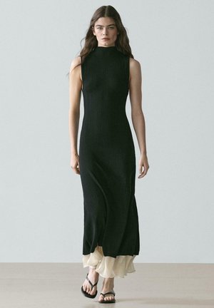 Donna con capelli lunghi e scuri che cammina, indossa un vestito maxi nero senza maniche a coste con orlo beige arricciato e sandali neri.