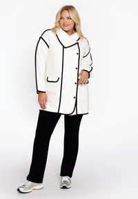 Witte jas met zwarte piping, grote kraag en knopen. Gestyled met zwarte broek en zilveren sneakers. Soepele textuur en gestructureerd ontwerp.