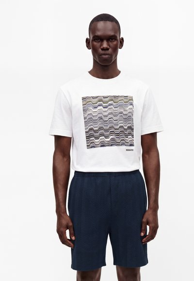 Homme portant un t-shirt blanc à manches courtes avec un motif carré en zigzag abstrait et un short texturé bleu marine, debout sur fond blanc.
