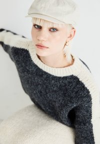 Maglione lavorato a maglia con strisce grigio scuro e crema, tessuto testurizzato, collo a costine e cuciture sulle spalle. Il modello indossa un cappello chiaro.