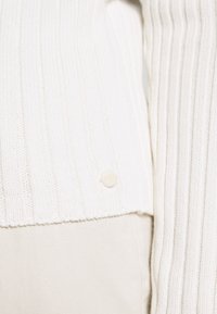 Cardigan bianco con coste, caratterizzato da un dettaglio con bottoni all'orlo, realizzato in un tessuto morbido con un'apparenza leggermente texturizzata.