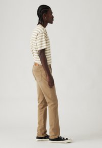 Chemise à manches courtes rayée en crème et beige, associée à un pantalon droit beige. Des baskets basses noires complètent la tenue.