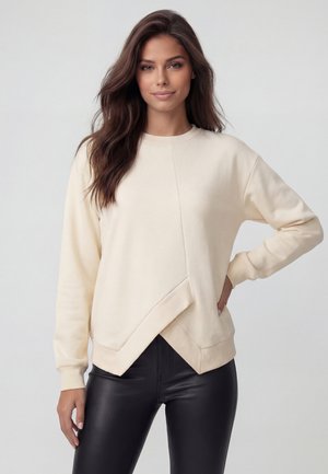 Frau mit langen braunen Haaren, die einen cremefarbenen Sweatshirt mit asymmetrischem Saum und schwarze Lederhosen trägt, steht mit einer Hand in der Hüfte.