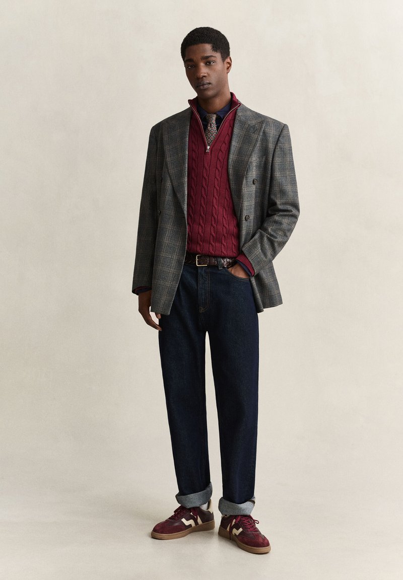 Man wearing gray checkered blazer, red cable-knit quarter-zip sweater, patterned tie, dark jeans rolled at cuffs, and red sneakers.