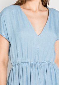 Sininen farkkumekko, jossa on syvä V-kaula-aukko, lyhyet hihat ja joustava vyötärö, joka johtaa kootun, valuvaan helmakeskukseen.