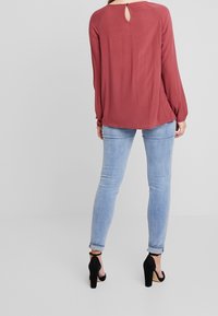 Långärmad blus i rostfärg med en nyckelhålsknäppning bak. Kombinerad med ljusblå jeans och svarta högklackade skor, vilket visar en avslappnad outfit.