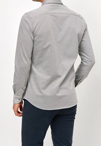 Mężczyzna w jasnoszarej, długiej koszuli z guzikami i ciemnych granatowych spodniach, przedstawiony od tyłu na jednolitym białym tle.