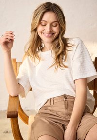 Femme souriante aux longs cheveux blonds portant un haut blanc texturé et un short beige, assise détendue sur une chaise en bois.