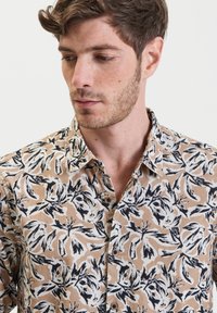Camicetta a maniche corte in beige chiaro con un motivo floreale nero. Realizzata in tessuto testurizzato, presenta un colletto a camicia e chiusura con bottoni frontali.