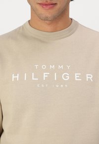 Sudadera de algodón beige con cuello redondo, textura suave y texto impreso en blanco: "TOMMY HILFIGER EST. 1985" en el frente.