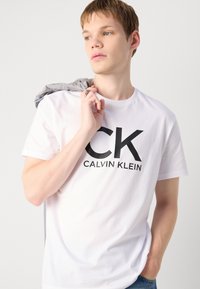 Witte katoenen t-shirt met een groot zwart "CK" logo en de tekst "CALVIN KLEIN". Model houdt een grijze kledingstuk over één schouder.