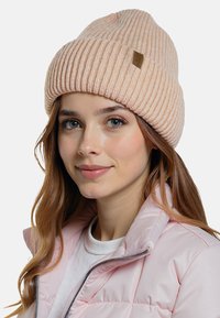 Vaaleanpunainen neulottu pipo, jossa on ribbitettu pinta ja käännetty reunus, jossa on pieni ruskea nahkainen logotarra. Pipoa on käytetty vaaleanpunaisen pörröisen takin kanssa.