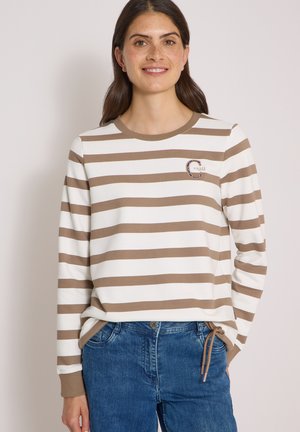 Femme aux longs cheveux bruns portant un t-shirt à manches longues rayé marron et blanc et un jean bleu, debout avec une main dans la poche.