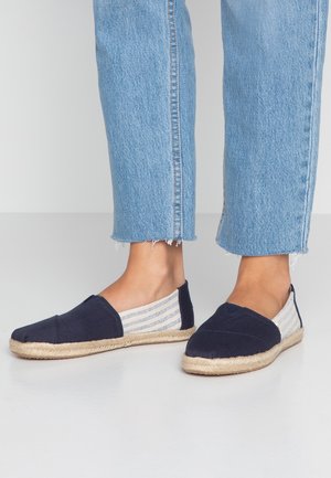 Granatowe espadryle typu slip-on z paskowanymi bokami z tkaniny, noszone z jasnoniebieskimi przyciętymi dżinsami na bosych stopach, stojące na szarej podłodze.