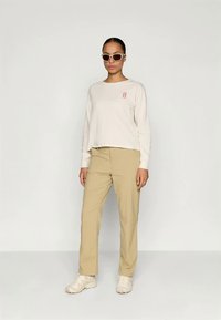 Long-sleeve cream top with red graphic, paired with beige pants and white sneakers. Model wearing white sunglasses, standing on a neutral background.