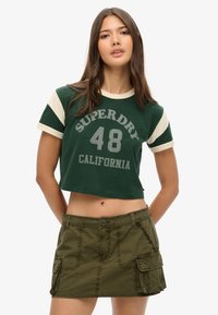 Groen cropped T-shirt met witte accenten, voorzien van een grafische opdruk met "SUPERDRY 48 CALIFORNIA". Gecombineerd met een olijfgroene rok met cargozakken.