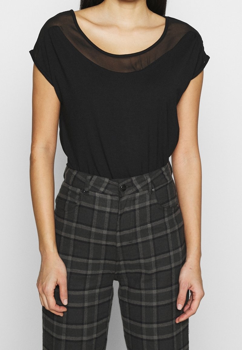 Femme portant un haut noir à manches courtes avec un décolleté transparent et un pantalon à carreaux gris taille haute, debout devant un fond uni.