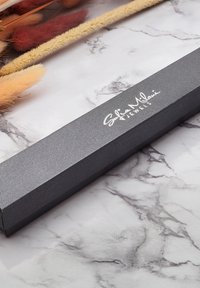 Svart texturerad smyckeslåda med rektangulär form och en blank logotyp. Placerad på en marmor yta med mjuka dekorativa element.