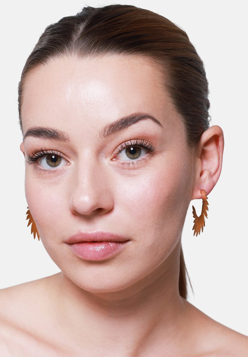 Bronzen blaadjesvormige oorbellen met ingewikkelde uitsparingen, hangend aan haken, gedragen door een model met een gladde, natuurlijke huid en minimale make-up.