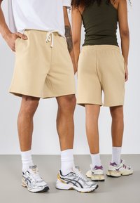 Shorts en coton beiges avec une taille élastique et un cordon de serrage, associés à des baskets de sport. Arrière-plan neutre, montrant l'ensemble de la tenue.