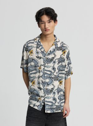 Młody mężczyzna w białej koszuli z krótkim rękawem z niebieskim smokiem i żółtymi kwiatowymi wzorami, stojący na jednolitym jasnym tle.