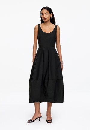 Mujer con vestido midi negro sin mangas con falda plisada, tacones negros de punta abierta, pendientes de perlas y una pulsera translúcida, de pie.