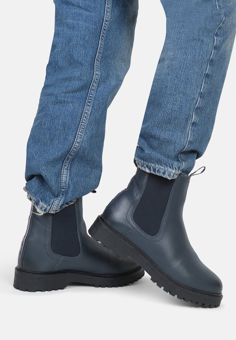 Bottines Chelsea bleu foncé en cuir, avec des panneaux latéraux élastiques et des semelles en caoutchouc texturé, portées avec un jean bleu clair retroussé.