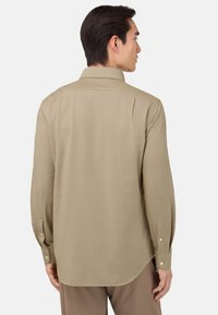 Camisa de manga larga en tejido beis claro con cuello puntiagudo, puños con botón y dobladillo redondeado, que presenta una textura suave y costuras sutiles.