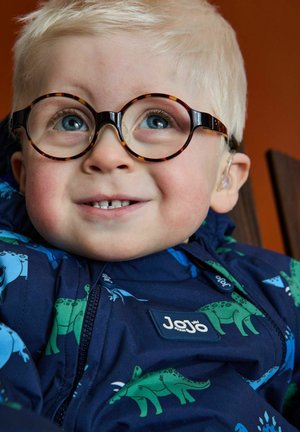 Jūrinės spalvos švarkas su dinozaurų raštais žalia ir mėlyna, apvalūs vėžlio lukšto akiniai, šviesiai blondinės plaukai ir ausinės prietaisas.