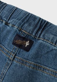 Fascia in denim con texture arricciata. Presenta un'etichetta blu scuro con la scritta "Adventures" e un logo a forma di cactus in rilievo. Cuciture visibili.