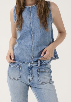 Frau trägt ärmelloses blaues Denim-Top mit rohem Saum und dazu passende hochtaillierte blaue Denim-Jeans mit großen Gesäßtaschen.