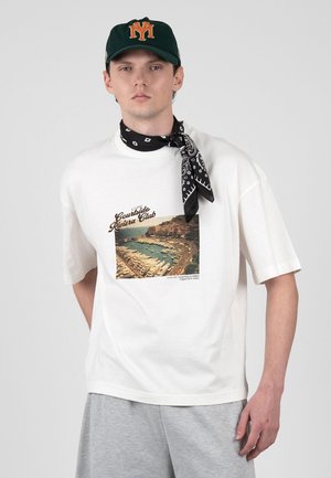 Ung mand iført hvid t-shirt med grafisk tryk af kystnær marina, grå shorts, sort bandana om halsen og mørkegrøn kasket med orange logo.
