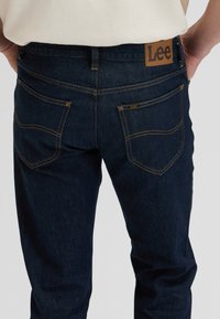 Άτομο που φοράει σκούρα μπλε τζιν Lee με ορατές πίσω τσέπες, κίτρινη ραφή και καφέ δερμάτινο σήμα της μάρκας στη μέση.