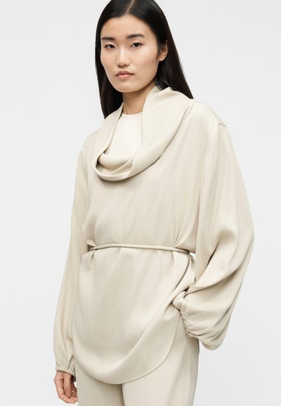 Μπεζ μακρυμάνικη μπλούζα με λαιμόκοψη τύπου ζιβάγκο, ελαστική μέση και μανίκια με σούρες. Μαλακό, λείο ύφασμα με άνετη εφαρμογή.