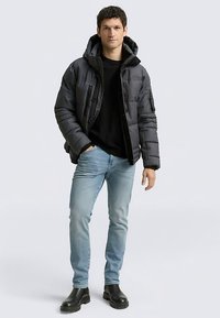 Čierna prešívaná bunda s kapucňou, prednými vreckami a zipsami; nosená cez čierne tričko a svetlomodré džínsy; doplnená čiernymi čižmami.