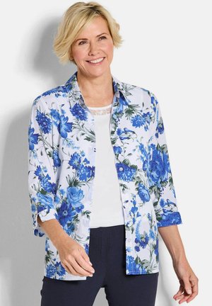 Lachende vrouw met kort blond haar, gekleed in een wit shirt onder een blauw-wit bloemenoverhemd en donkere broek.