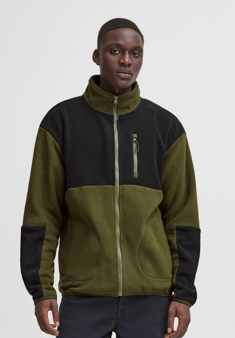Chaqueta de forro polar en verde oliva y negro con un alto cuello, cierre de cremallera, dos bolsillos frontales y un pequeño bolsillo en el pecho con cremallera.