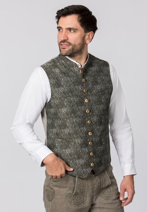 Grüner gemusterter Weste mit hohem Kragen, Holzknöpfen und braunem Paspel, getragen über einem weißen Hemd. Verfügt über zwei Fronttaschen.