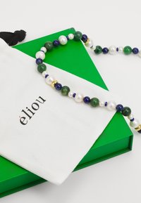 Bunte Perlenkette mit grünen Steinen, blauen Lapislazuli und weißen Perlen, auf einem weißen Beutel mit "élieou" und einer grünen Box platziert.